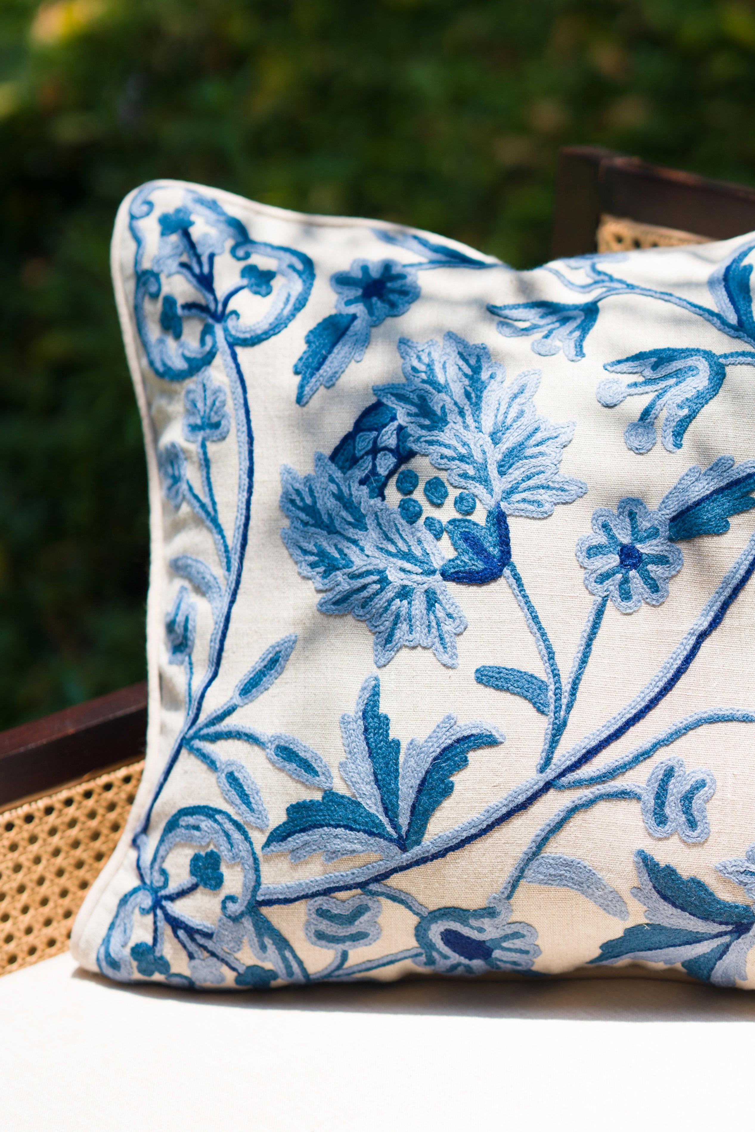 Blue crewel embroidered linen cushion styled outdoors among greenery.