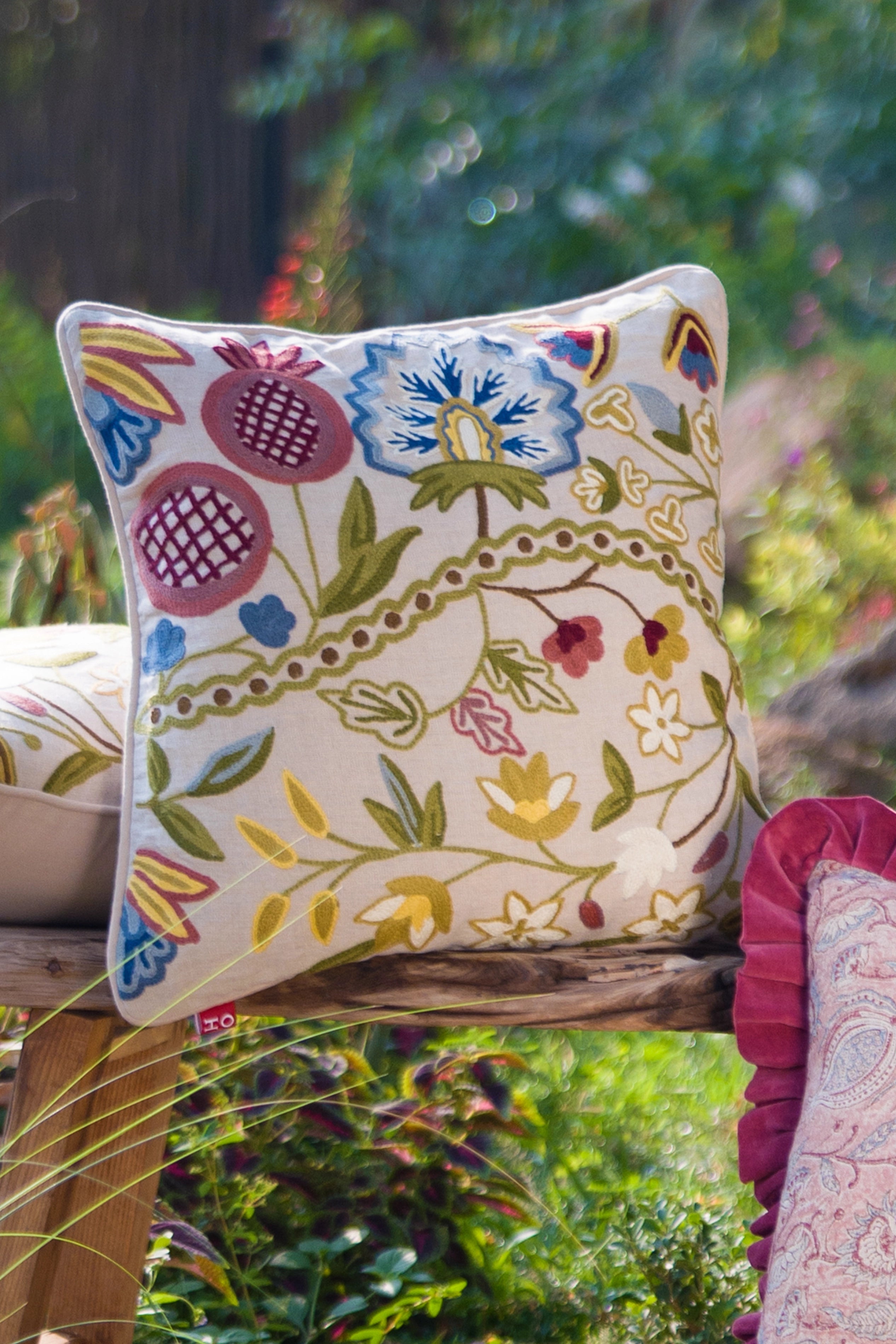 Square multicolour floral crewel embroidered linen cushion styled outdoors among greenery.