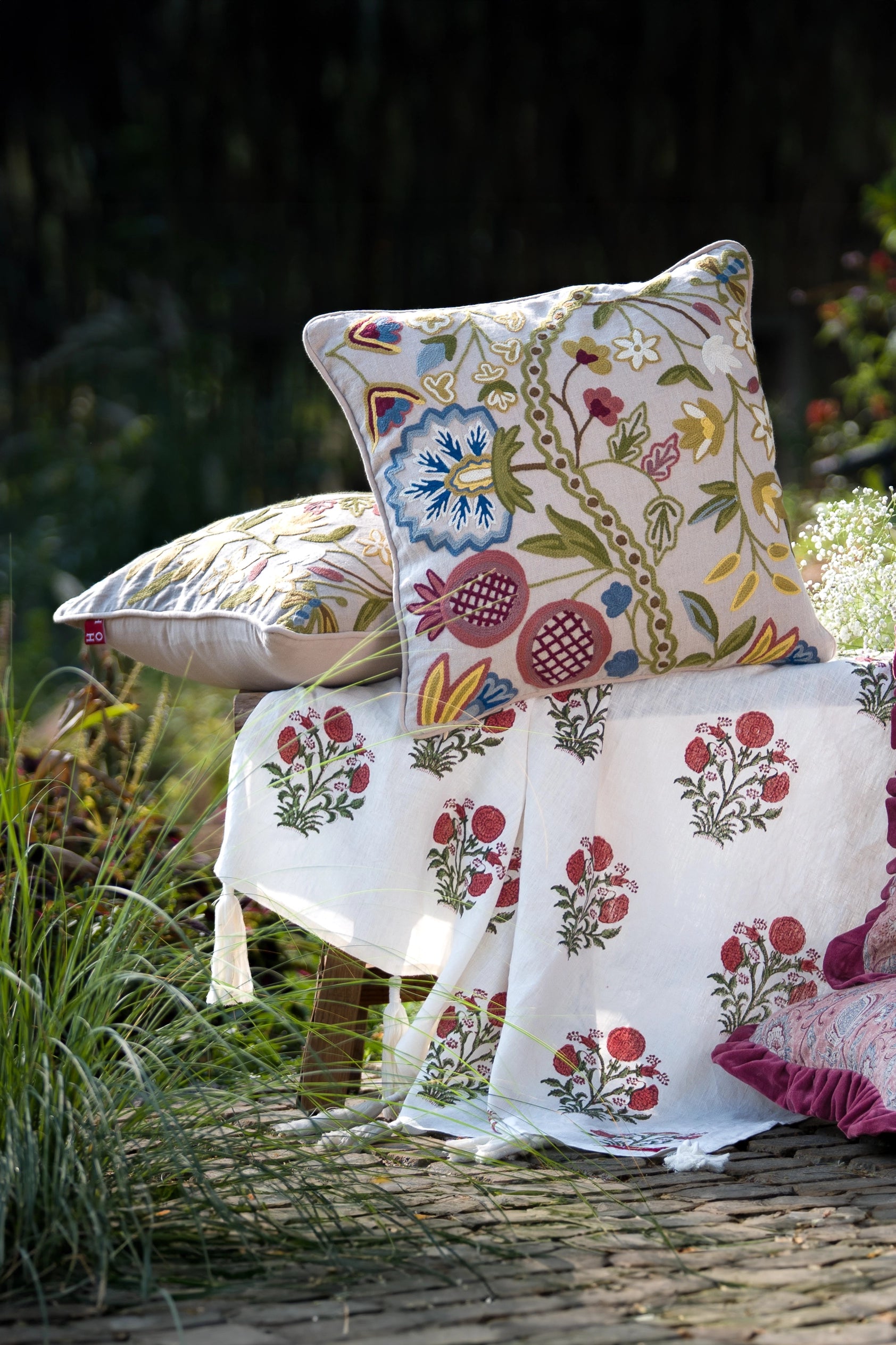 Square multicolour floral crewel embroidered linen cushion styled on outdoor seating with matching cushions.
