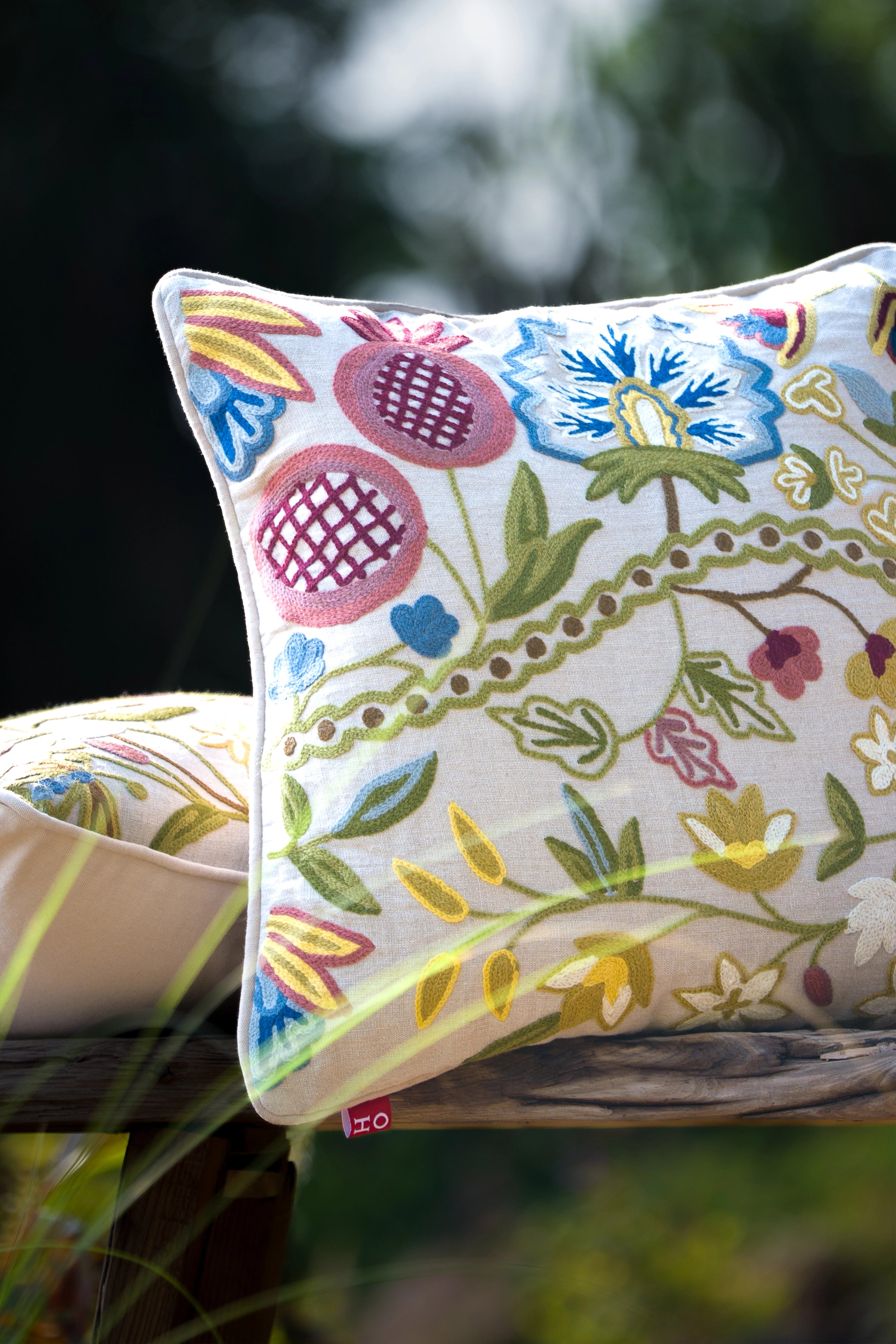Square multicolour floral crewel embroidered linen cushion styled outdoors among garden foliage.